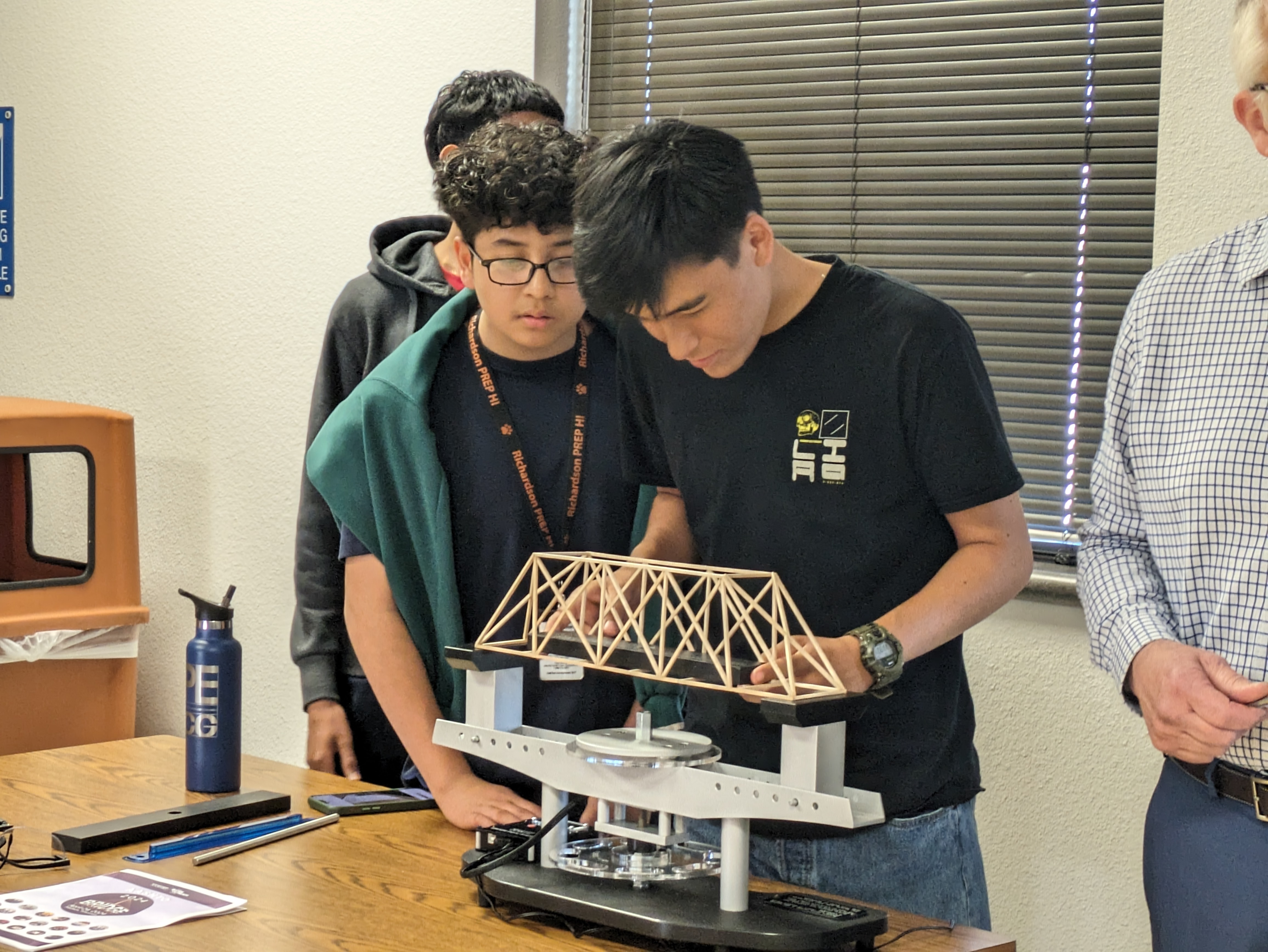 Students testing a small wooden bridge model on a load-bearing scale during an engineering class activity.