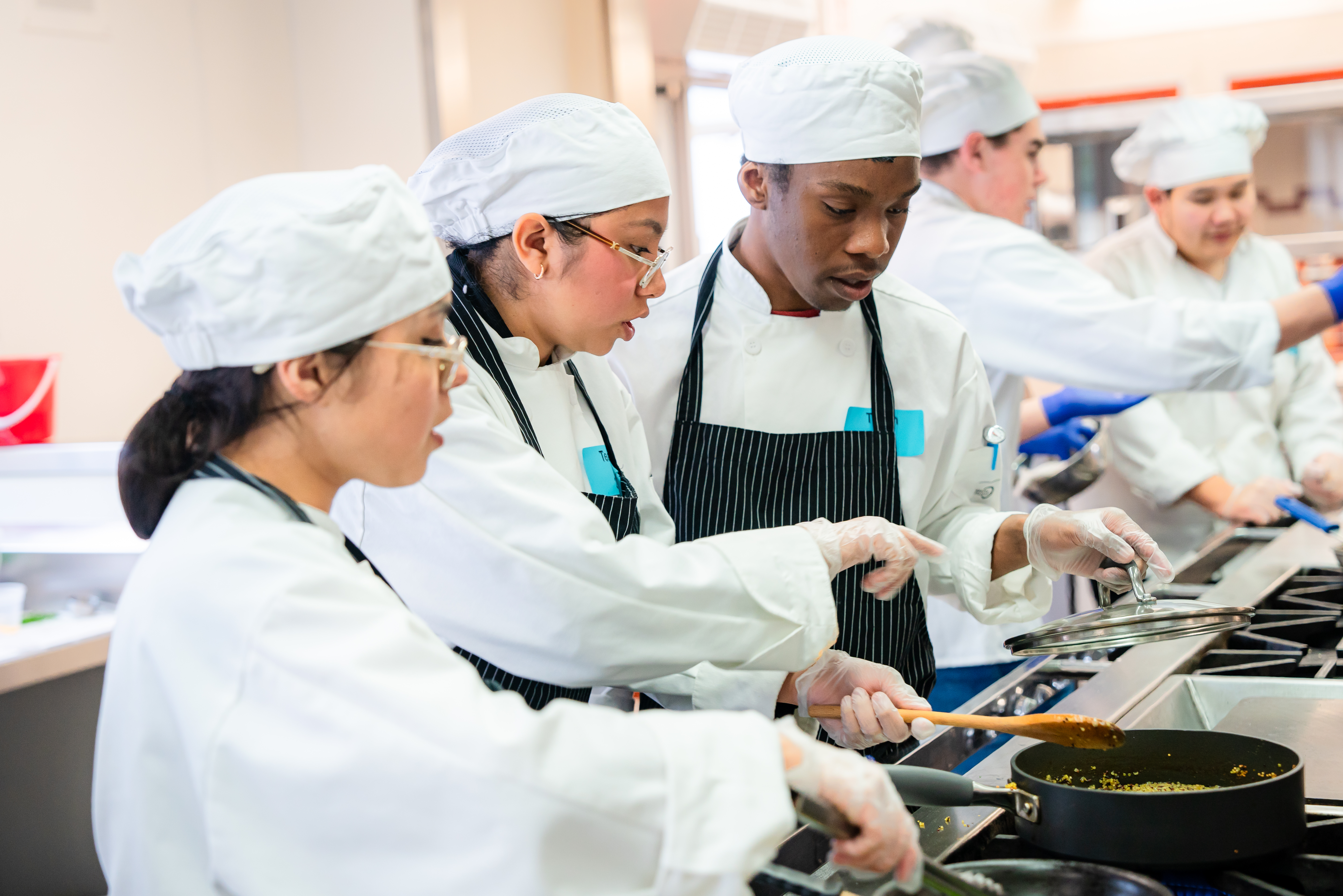 Culinary students in chef uniforms collaborating at a stovetop station while preparing a dish.