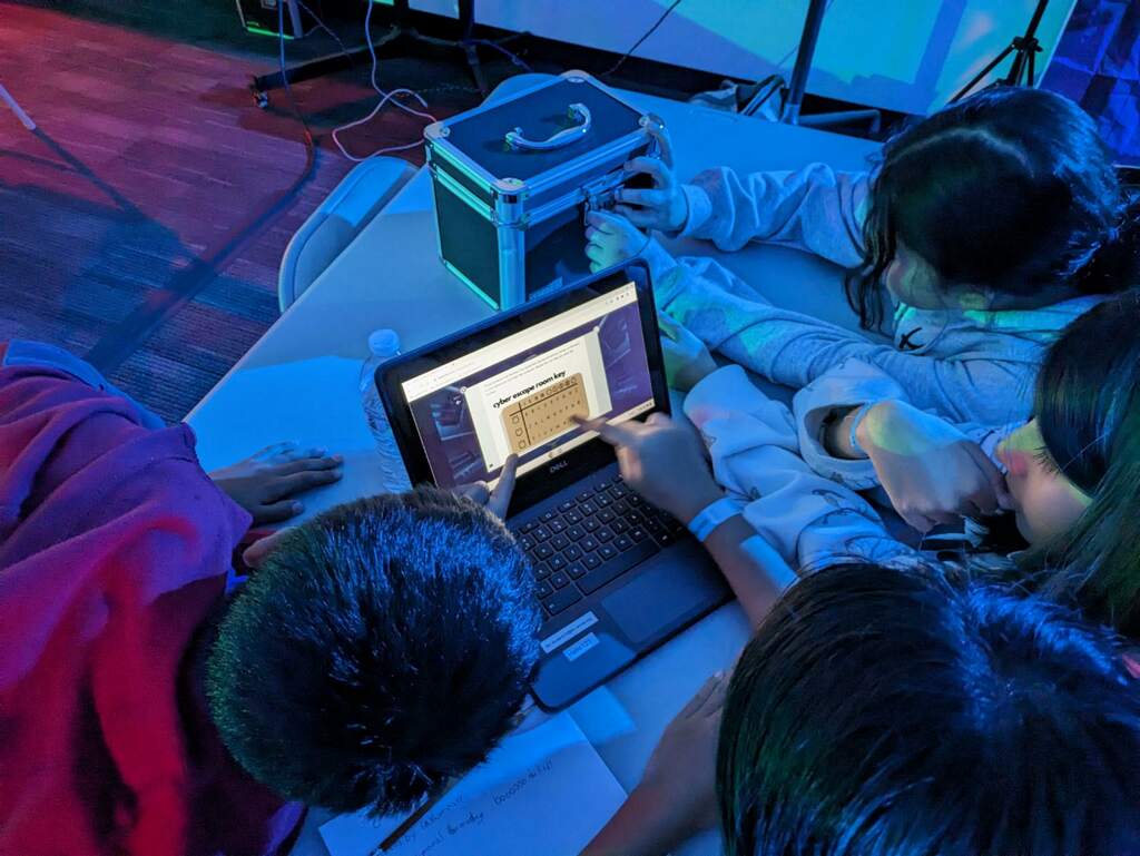 Group of students working together in a darkened room on a cybersecurity challenge displayed on a laptop.