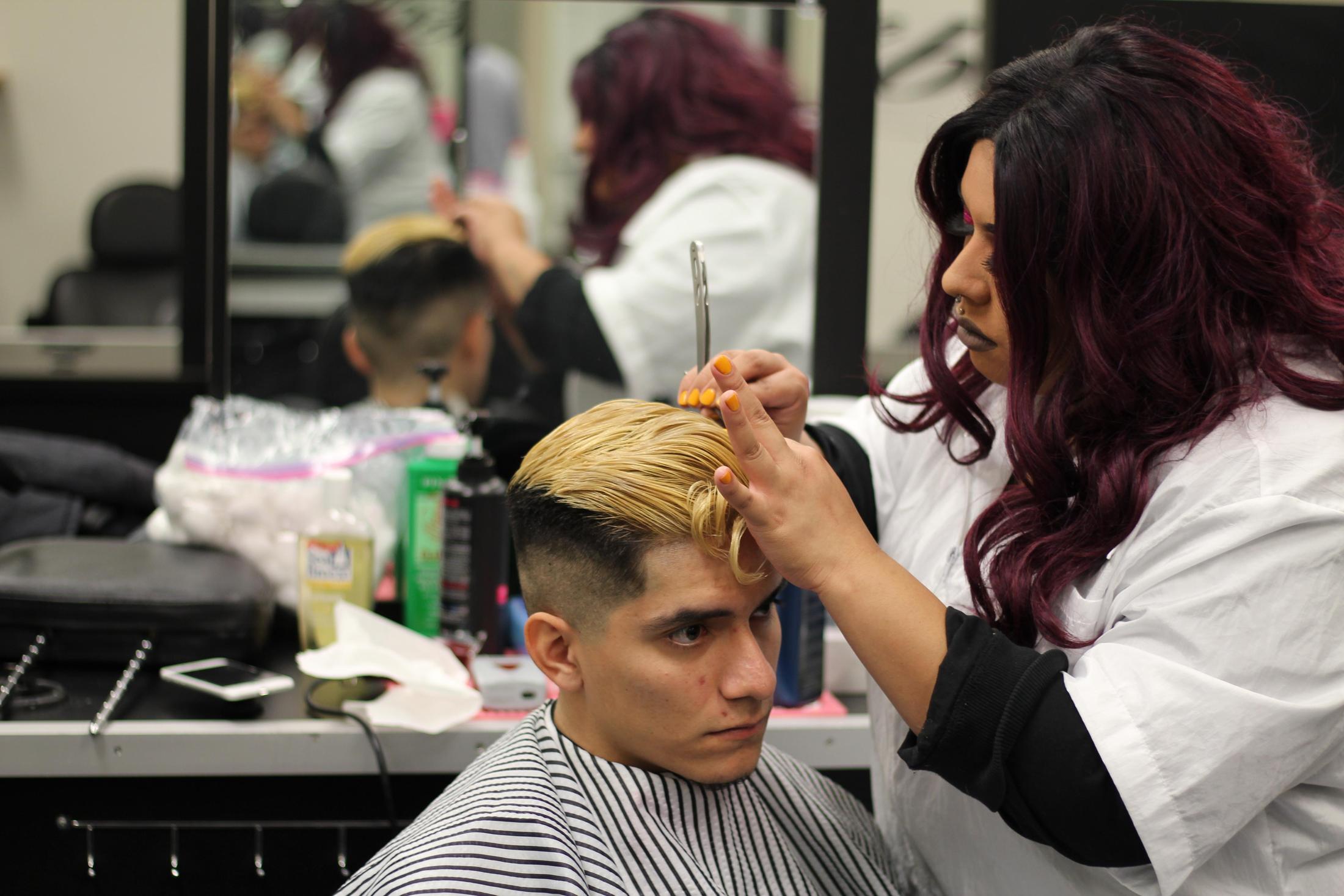 A cosmetology student trims the hair of a client with a blonde top and faded sides in a salon classroom.