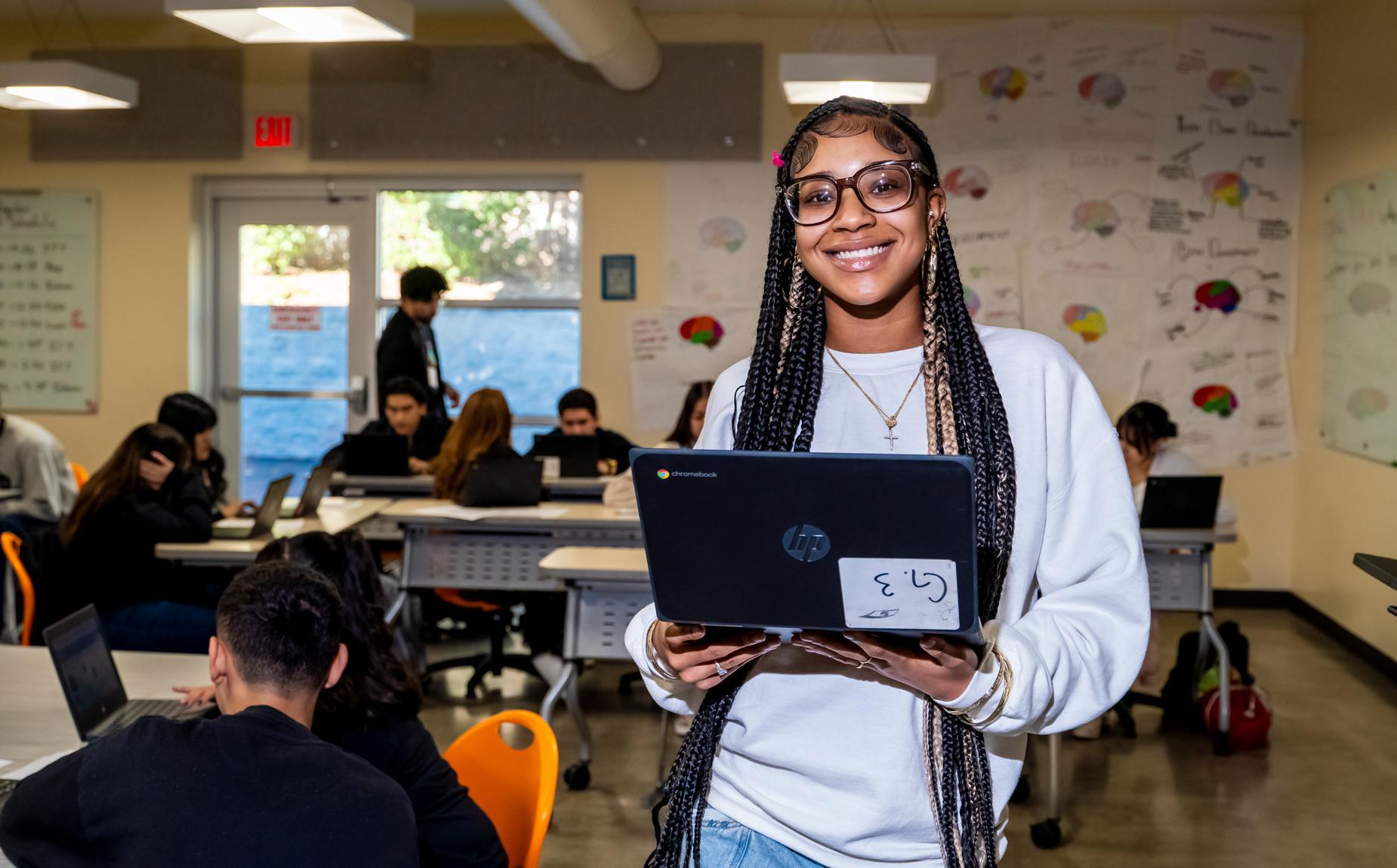 A student smiles while holding a Chromebook in a busy classroom where classmates work on laptops.